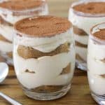 Four individual servings of tiramisu in clear glass jars are neatly arranged on a wooden table. Each jar contains layers of mascarpone cheese, ladyfingers, and cocoa powder. Two silver spoons are placed beside the jars.