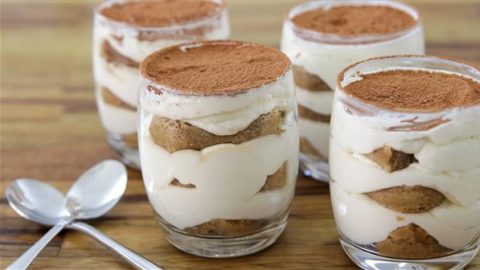 Four individual servings of tiramisu in clear glass jars are neatly arranged on a wooden table. Each jar contains layers of mascarpone cheese, ladyfingers, and cocoa powder. Two silver spoons are placed beside the jars.