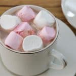 A white ceramic cup filled with hot chocolate is topped with pink and white marshmallows. The cup rests on a matching saucer placed on a wooden surface, with another saucer partially visible in the background.