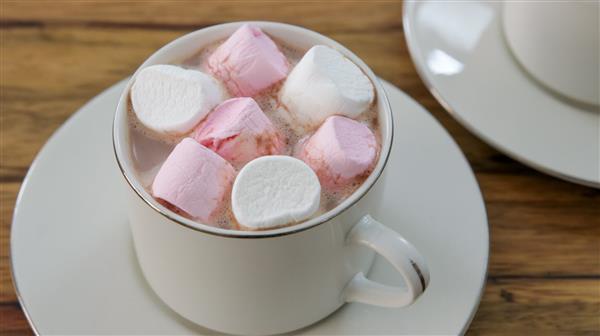 A white ceramic cup filled with hot chocolate is topped with pink and white marshmallows. The cup rests on a matching saucer placed on a wooden surface, with another saucer partially visible in the background.