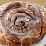 A close-up of a freshly baked cinnamon roll covered with a light drizzle of icing. The cinnamon roll has a golden-brown color and is placed on a white plate atop a wooden surface.