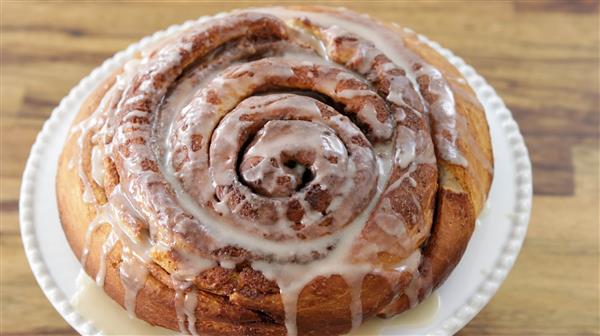 A close-up of a freshly baked cinnamon roll covered with a light drizzle of icing. The cinnamon roll has a golden-brown color and is placed on a white plate atop a wooden surface.