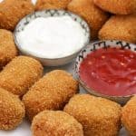 A plate of golden-brown, breaded chicken nuggets is arranged around two small bowls of dipping sauces: one with a white creamy sauce and the other with a red chili sauce. The plate is set on a wooden surface.