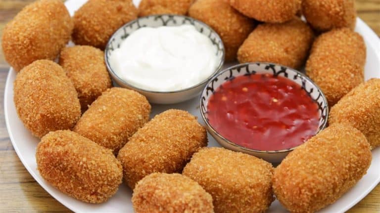 A plate of golden-brown, breaded chicken nuggets is arranged around two small bowls of dipping sauces: one with a white creamy sauce and the other with a red chili sauce. The plate is set on a wooden surface.