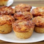 A white plate holds several savory muffins with a golden-brown, cheesy topping. They appear freshly baked, with a crusty exterior and small, round shape. A second plate with more muffins is partially visible in the background on a wooden surface.