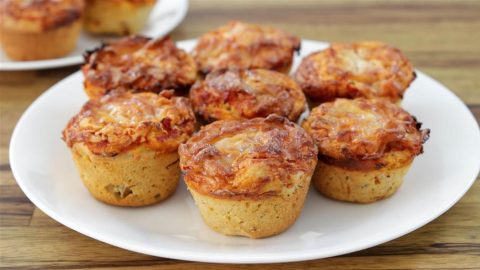 A white plate holds several savory muffins with a golden-brown, cheesy topping. They appear freshly baked, with a crusty exterior and small, round shape. A second plate with more muffins is partially visible in the background on a wooden surface.