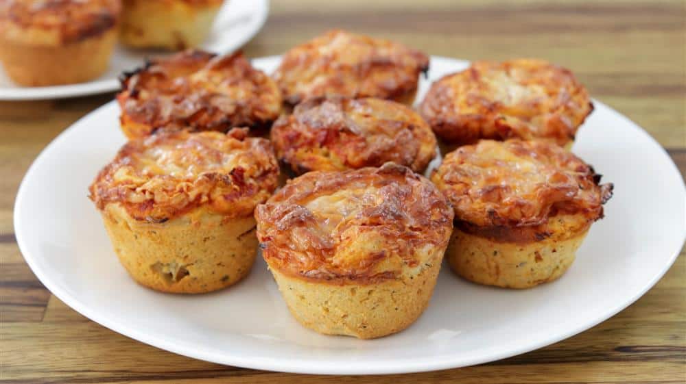 A white plate holds several savory muffins with a golden-brown, cheesy topping. They appear freshly baked, with a crusty exterior and small, round shape. A second plate with more muffins is partially visible in the background on a wooden surface.