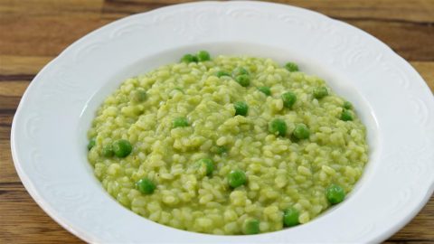 A white bowl filled with a creamy green pea risotto is placed on a wooden surface. The risotto features cooked rice mixed with bright green peas, all coated in a smooth green sauce, giving the dish a vibrant and appetizing appearance.