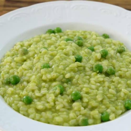 A white bowl filled with a creamy green pea risotto is placed on a wooden surface. The risotto features cooked rice mixed with bright green peas, all coated in a smooth green sauce, giving the dish a vibrant and appetizing appearance.