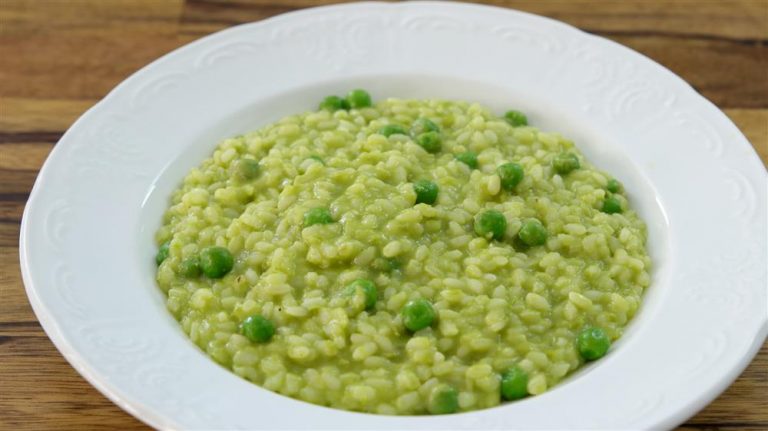 A white bowl filled with a creamy green pea risotto is placed on a wooden surface. The risotto features cooked rice mixed with bright green peas, all coated in a smooth green sauce, giving the dish a vibrant and appetizing appearance.