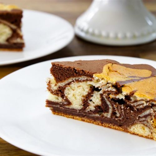 A slice of marble cake, which features swirls of vanilla and chocolate layers, sits on a white plate placed on a wooden table. In the background, another plate with a similar slice and a cake stand can be seen. A gold fork rests beside the plate.