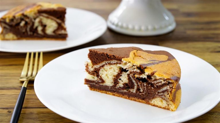 A slice of marble cake, which features swirls of vanilla and chocolate layers, sits on a white plate placed on a wooden table. In the background, another plate with a similar slice and a cake stand can be seen. A gold fork rests beside the plate.