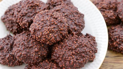 A white plate full of chocolate-covered quinoa clusters. The clusters are round and rough-textured, with visible quinoa grains coated in dark chocolate. Another group of clusters is partially visible in the background.