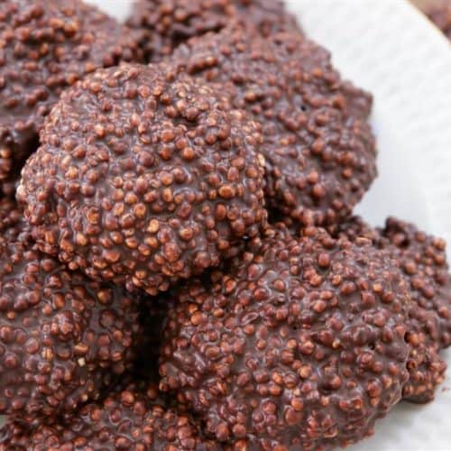 A white plate full of chocolate-covered quinoa clusters. The clusters are round and rough-textured, with visible quinoa grains coated in dark chocolate. Another group of clusters is partially visible in the background.