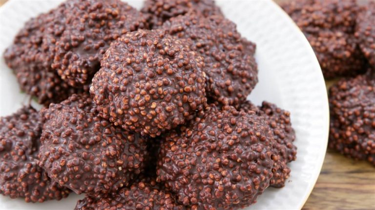 A white plate full of chocolate-covered quinoa clusters. The clusters are round and rough-textured, with visible quinoa grains coated in dark chocolate. Another group of clusters is partially visible in the background.