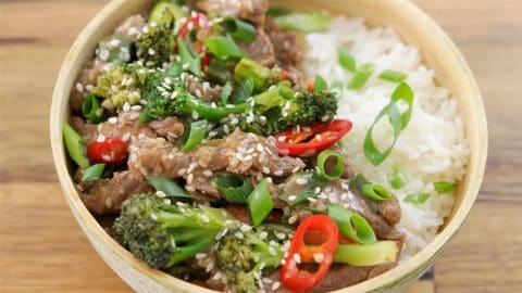 A bowl of white rice topped with a stir-fry of beef strips, broccoli florets, sliced chili peppers, green onions, and sesame seeds. The dish is colorful, with vibrant green broccoli, red chili slices, and a sprinkle of sesame seeds for garnish.