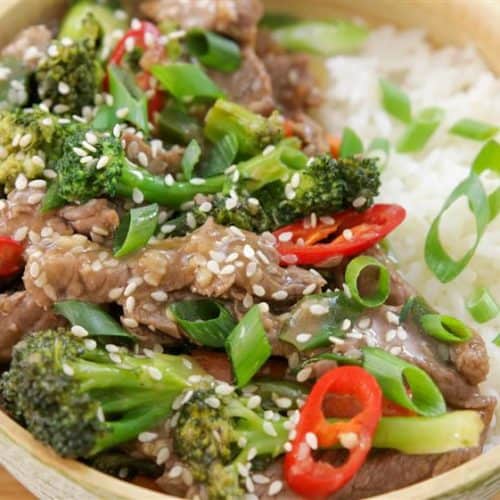 A bowl of white rice topped with a stir-fry of beef strips, broccoli florets, sliced chili peppers, green onions, and sesame seeds. The dish is colorful, with vibrant green broccoli, red chili slices, and a sprinkle of sesame seeds for garnish.