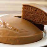 A chocolate cake with a smooth, glossy icing sits on a white plate. A slice has been cut and is being lifted from the cake, revealing its rich and moist interior. The background has a wooden texture.