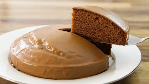 A chocolate cake with a smooth, glossy icing sits on a white plate. A slice has been cut and is being lifted from the cake, revealing its rich and moist interior. The background has a wooden texture.