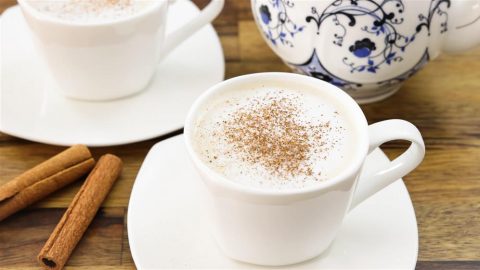 A white cup filled with frothy coffee topped with a sprinkle of cinnamon sits on a white saucer. Nearby, two cinnamon sticks and another cup with a similar setup are placed on a wooden surface. In the background, a decorative teapot is partially visible.