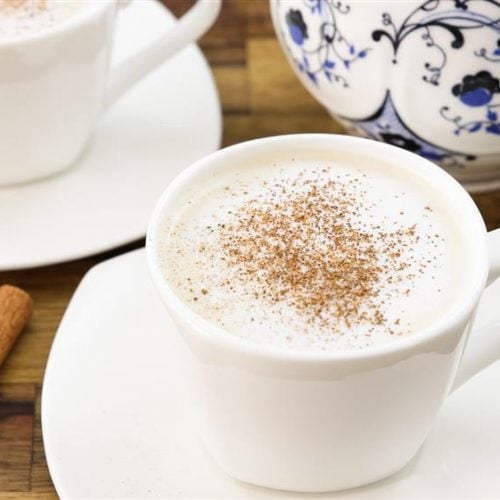 A white cup filled with frothy coffee topped with a sprinkle of cinnamon sits on a white saucer. Nearby, two cinnamon sticks and another cup with a similar setup are placed on a wooden surface. In the background, a decorative teapot is partially visible.