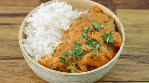 A bowl with half filled with white rice and half filled with a creamy, orange-colored curry garnished with chopped cilantro. The background is a wooden surface.