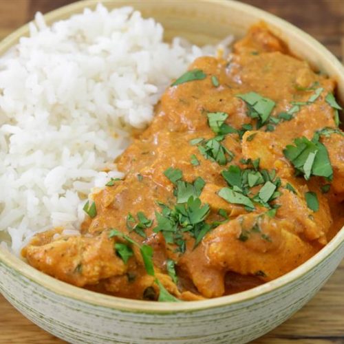 A bowl with half filled with white rice and half filled with a creamy, orange-colored curry garnished with chopped cilantro. The background is a wooden surface.