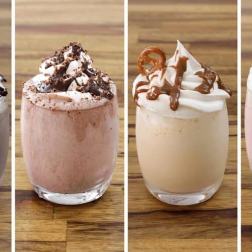 Four gourmet milkshakes are presented in a row. From left to right: a chocolate shake with whipped cream and cookie crumbles, a chocolate shake with whipped cream and chocolate shavings, a vanilla caramel shake with whipped cream and caramel drizzle, and a strawberry shake with whipped cream and a strawberry on top. They are displayed on a wooden table.