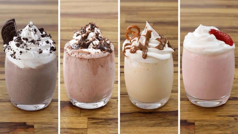 Four gourmet milkshakes are presented in a row. From left to right: a chocolate shake with whipped cream and cookie crumbles, a chocolate shake with whipped cream and chocolate shavings, a vanilla caramel shake with whipped cream and caramel drizzle, and a strawberry shake with whipped cream and a strawberry on top. They are displayed on a wooden table.