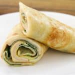 A white plate holds two neatly rolled chicken wraps. The wraps are partially cut to reveal slices of chicken, spinach, and a creamy spread inside, all encased in a toasted flatbread. The background features a brown wooden surface.