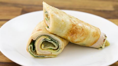 A white plate holds two neatly rolled chicken wraps. The wraps are partially cut to reveal slices of chicken, spinach, and a creamy spread inside, all encased in a toasted flatbread. The background features a brown wooden surface.