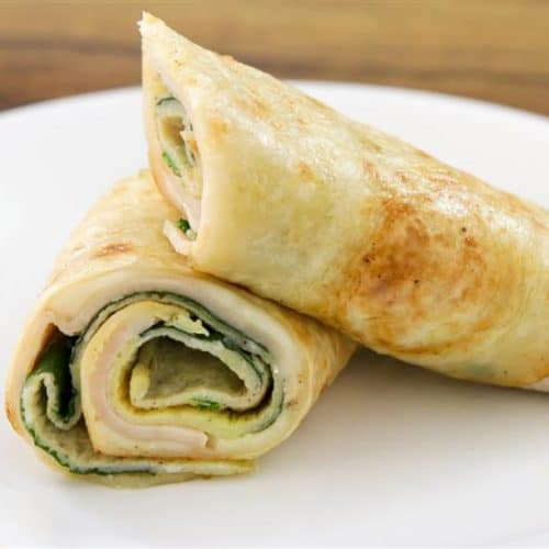 A white plate holds two neatly rolled chicken wraps. The wraps are partially cut to reveal slices of chicken, spinach, and a creamy spread inside, all encased in a toasted flatbread. The background features a brown wooden surface.