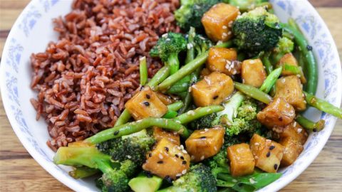 A plate featuring a serving of red rice on the left and a colorful stir-fry on the right, which includes tofu cubes, broccoli, green beans, sesame seeds, and a savory sauce, all arranged in a white dish with a blue floral rim.