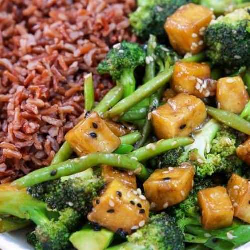 A plate featuring a serving of red rice on the left and a colorful stir-fry on the right, which includes tofu cubes, broccoli, green beans, sesame seeds, and a savory sauce, all arranged in a white dish with a blue floral rim.