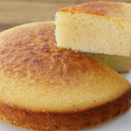 A hand holds a spatula lifting a slice of golden-brown cornbread from a round, evenly baked cornbread sitting on a white plate. The background features a wooden surface, slightly blurred.