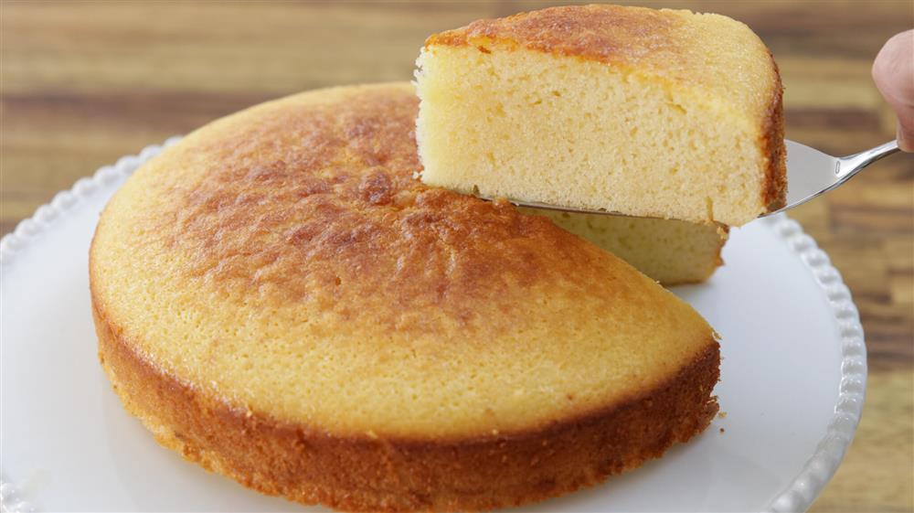 A hand holds a spatula lifting a slice of golden-brown cornbread from a round, evenly baked cornbread sitting on a white plate. The background features a wooden surface, slightly blurred.