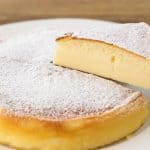 A fluffy, round Japanese cheesecake dusted with powdered sugar sits on a white plate. A slice is being lifted out with a utensil, revealing its soft and airy texture.