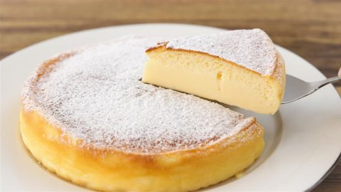 A fluffy, round Japanese cheesecake dusted with powdered sugar sits on a white plate. A slice is being lifted out with a utensil, revealing its soft and airy texture.