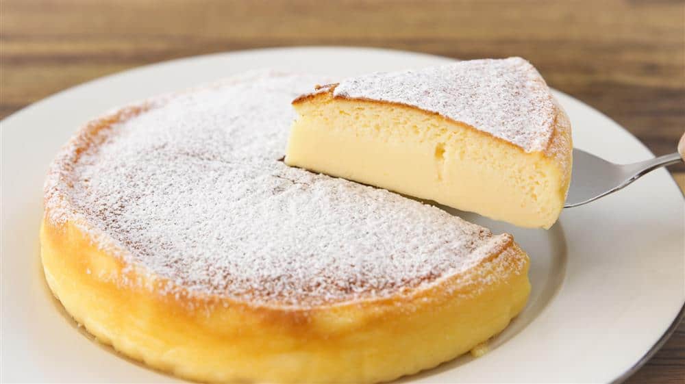 A fluffy, round Japanese cheesecake dusted with powdered sugar sits on a white plate. A slice is being lifted out with a utensil, revealing its soft and airy texture.