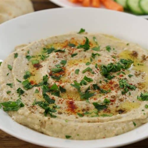 A white plate filled with creamy hummus topped with olive oil, chopped parsley, and a sprinkle of paprika is set on a wooden surface. In the background, there are slices of pita bread and a plate with sliced cucumbers and carrots.