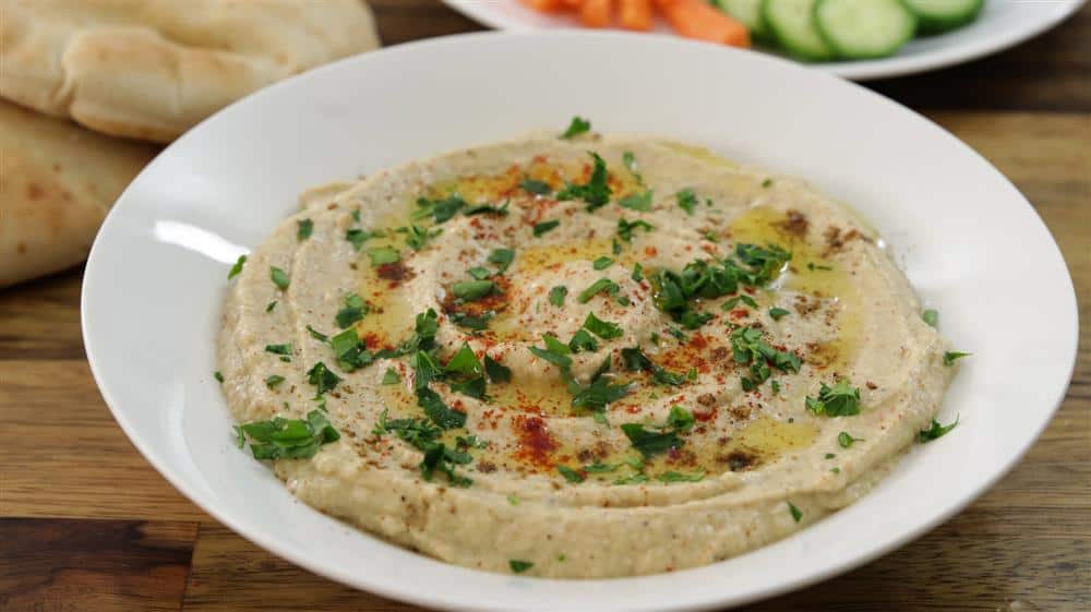 A white plate filled with creamy hummus topped with olive oil, chopped parsley, and a sprinkle of paprika is set on a wooden surface. In the background, there are slices of pita bread and a plate with sliced cucumbers and carrots.