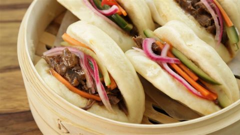 A close-up of a bamboo steamer containing three steamed bao buns filled with shredded beef, pickled red onions, julienned carrots, and cucumber sticks. The buns are soft and fluffy, and the ingredients look fresh and colorful.