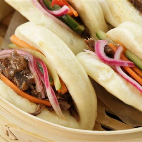 A close-up of a bamboo steamer containing three steamed bao buns filled with shredded beef, pickled red onions, julienned carrots, and cucumber sticks. The buns are soft and fluffy, and the ingredients look fresh and colorful.