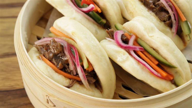 A close-up of a bamboo steamer containing three steamed bao buns filled with shredded beef, pickled red onions, julienned carrots, and cucumber sticks. The buns are soft and fluffy, and the ingredients look fresh and colorful.