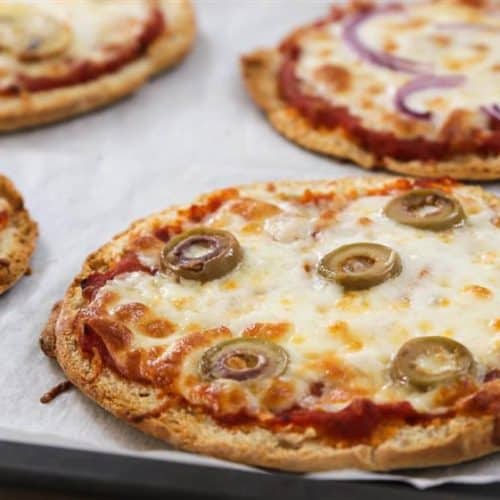 Four small pizzas with melted cheese, tomato sauce, and various toppings, including green olives and red onion slices, are placed on a baking sheet lined with parchment paper. The pizzas appear to be freshly baked and have a crispy crust.