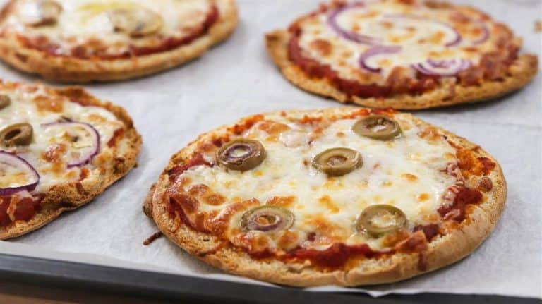 Four small pizzas with melted cheese, tomato sauce, and various toppings, including green olives and red onion slices, are placed on a baking sheet lined with parchment paper. The pizzas appear to be freshly baked and have a crispy crust.