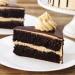 A slice of layered chocolate cake with a creamy filling and topped with a swirl of frosting on a white plate. Another similar slice and the rest of the cake on a white cake stand are visible in the background. A gold fork is placed next to the plate.