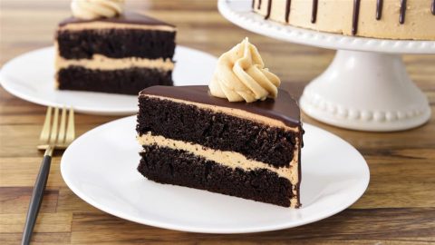 A slice of layered chocolate cake with a creamy filling and topped with a swirl of frosting on a white plate. Another similar slice and the rest of the cake on a white cake stand are visible in the background. A gold fork is placed next to the plate.