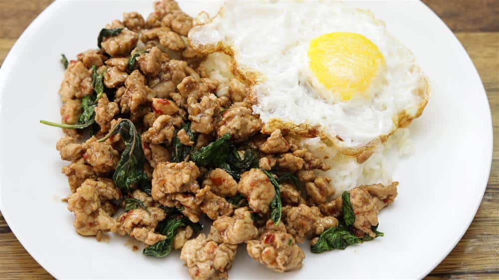 A plate of Thai basil chicken stir-fry served with a fried egg on steamed white rice. The dish showcases minced chicken mixed with basil leaves and red chili slices, next to a sunny-side-up egg with a runny yolk.