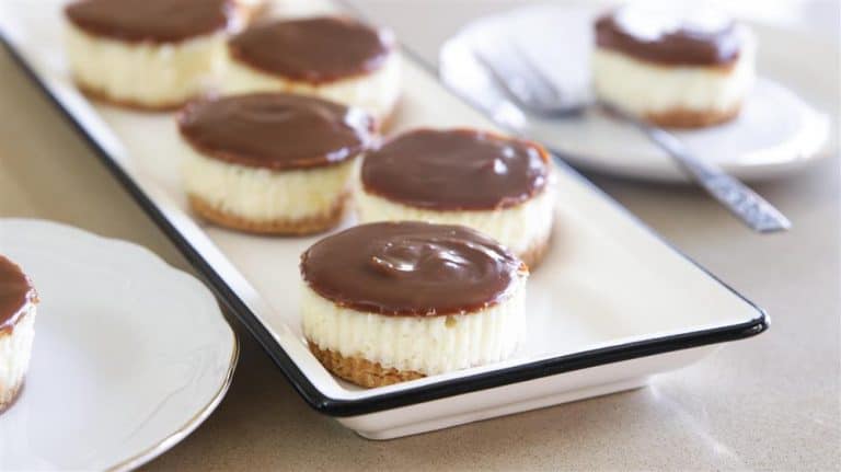 A close-up view of several small cheesecakes topped with a layer of chocolate ganache, neatly arranged on a white rectangular serving platter. A single cheesecake is placed separately on a white plate with a fork in the background.